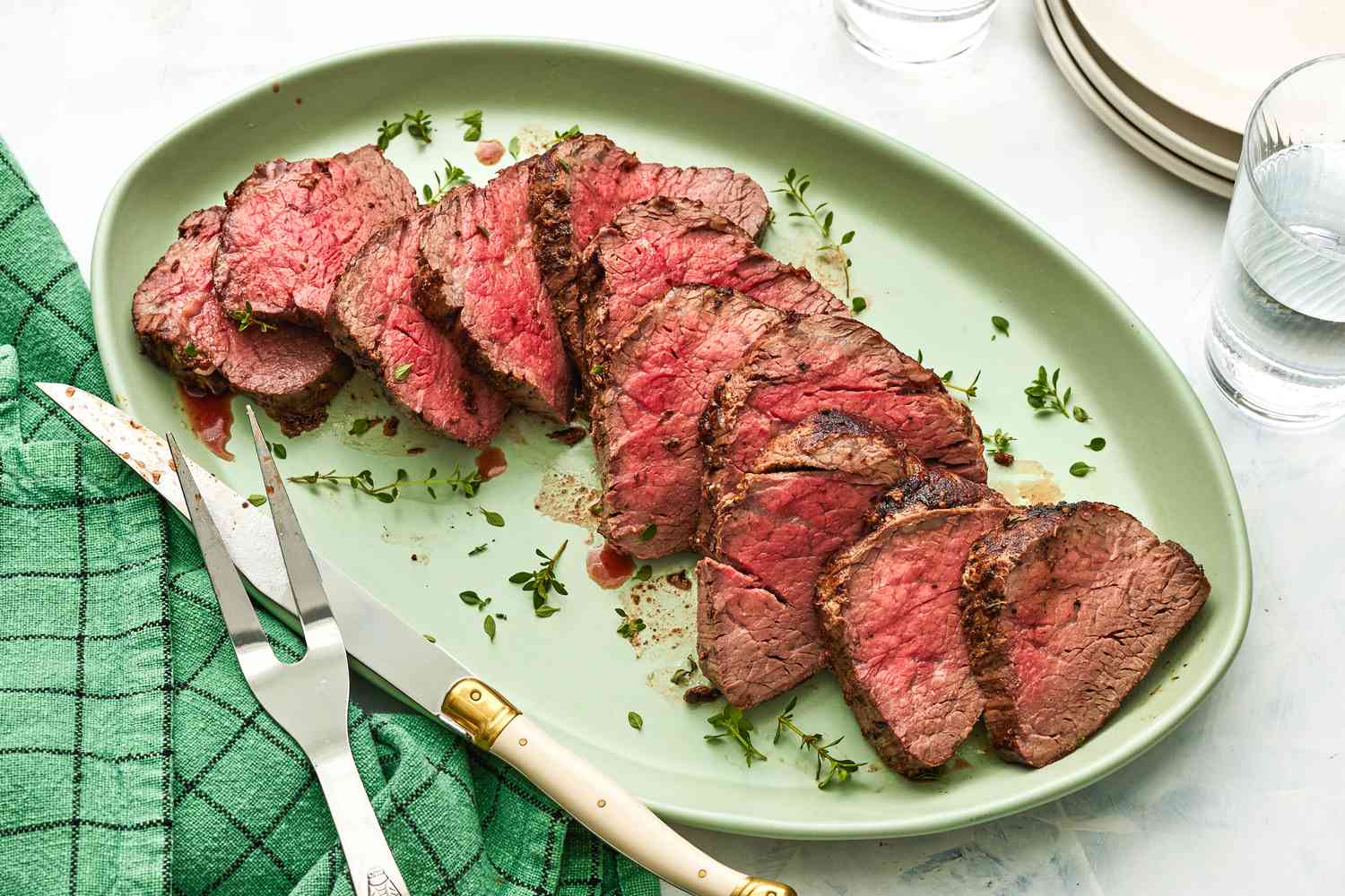 Sliced medium rare beef fillet steaks on green serving plate with fresh herbs carving knife and fork