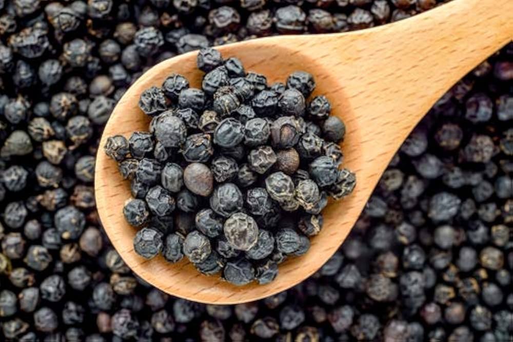 Whole dried black peppercorns on wooden spoon with scattered black pepper grains close-up for spice product photography