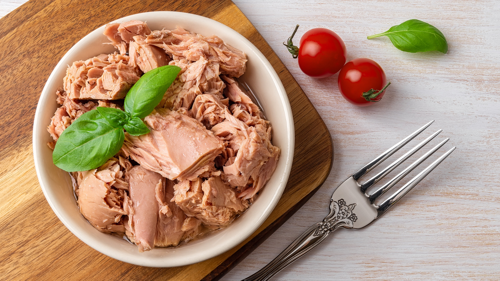 Top view of canned tuna chunks in white bowl with fresh basil leaves cherry tomatoes and fork on wooden board