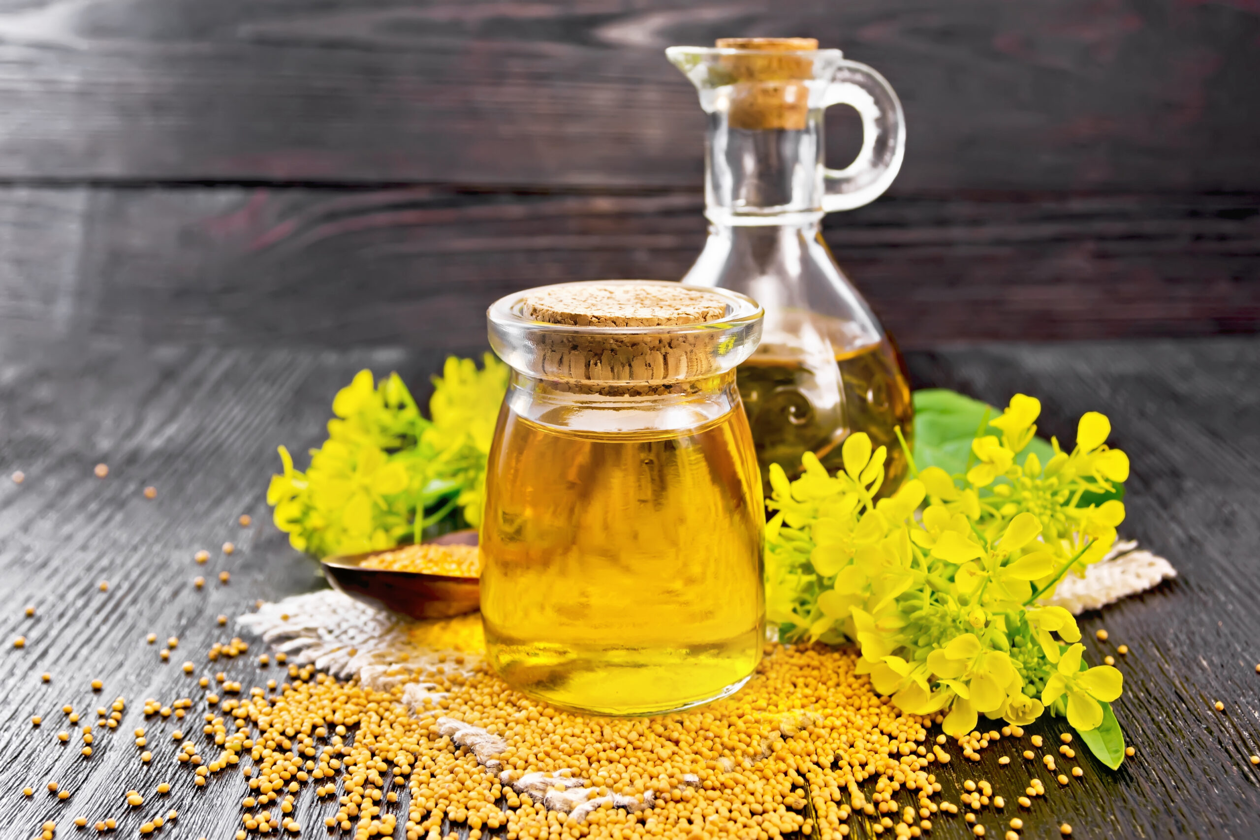 Mustard oil in a glass jar and decanter, mustard grains on a burlap napkin, flowers and leaves on wooden board background