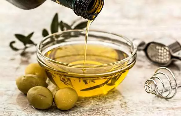 Golden extra virgin olive oil pouring from bottle into glass bowl with fresh green olives and olive branch on stone surface