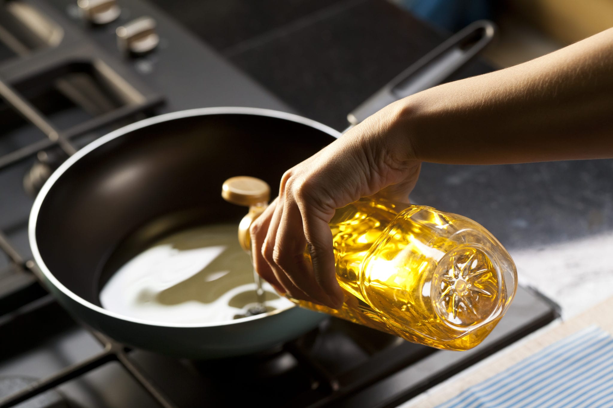 Hand pouring golden cooking oil from bottle into black frying pan on kitchen stove for food preparation photography